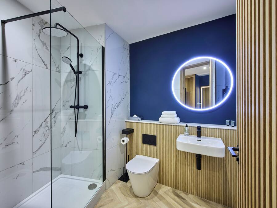 Un baño moderno con ducha envuelta en vidrio, inodoro blanco, tocador de madera con lavamanos y espejo con luz circular.
