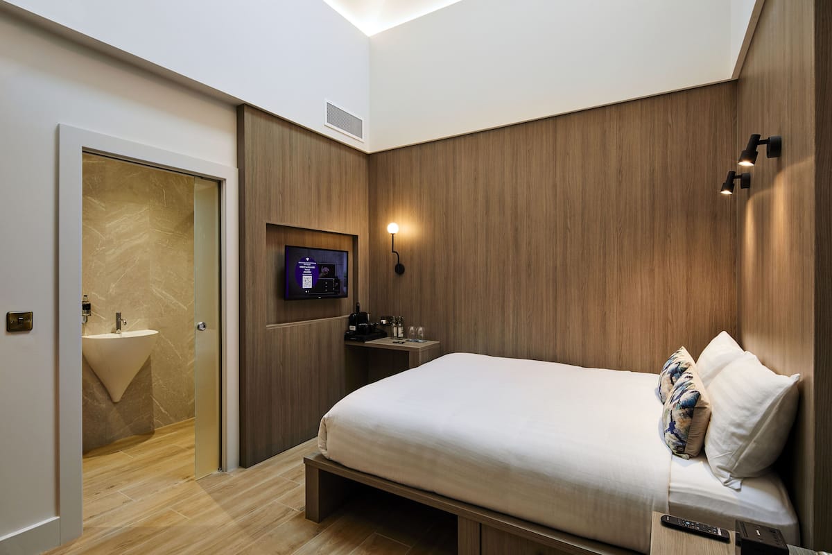 Una habitación de hotel moderna con cama, televisión, mesita de noche y baño con lavamanos y espejo.