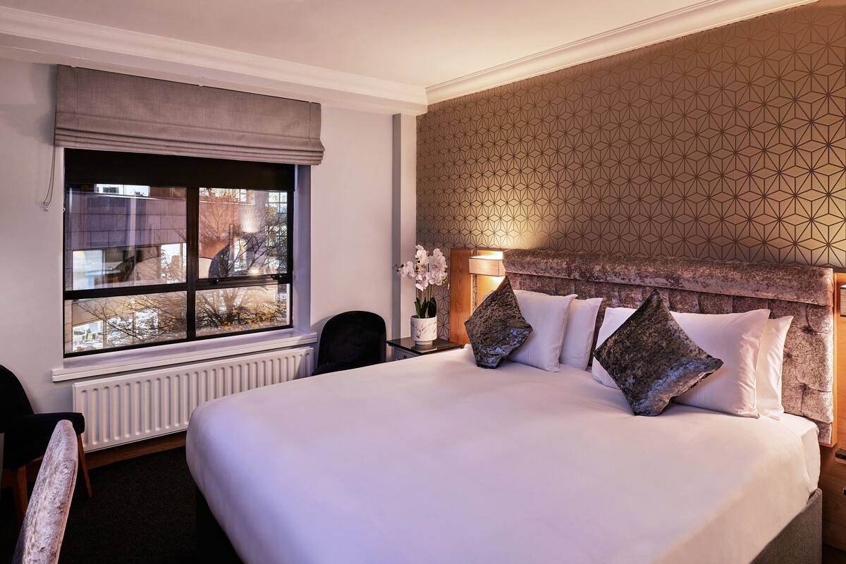 Habitación de hotel con una cama grande, una mesita de noche con lámpara, un ventanal con vistas y una pared con diseño.