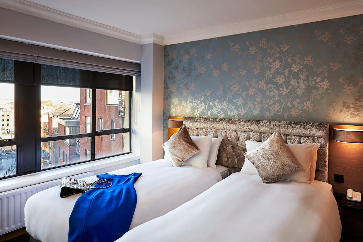 Una habitación de hotel con dos camas, vistas a la ciudad a través de amplias ventanas y un papel tapiz con motivos florales.