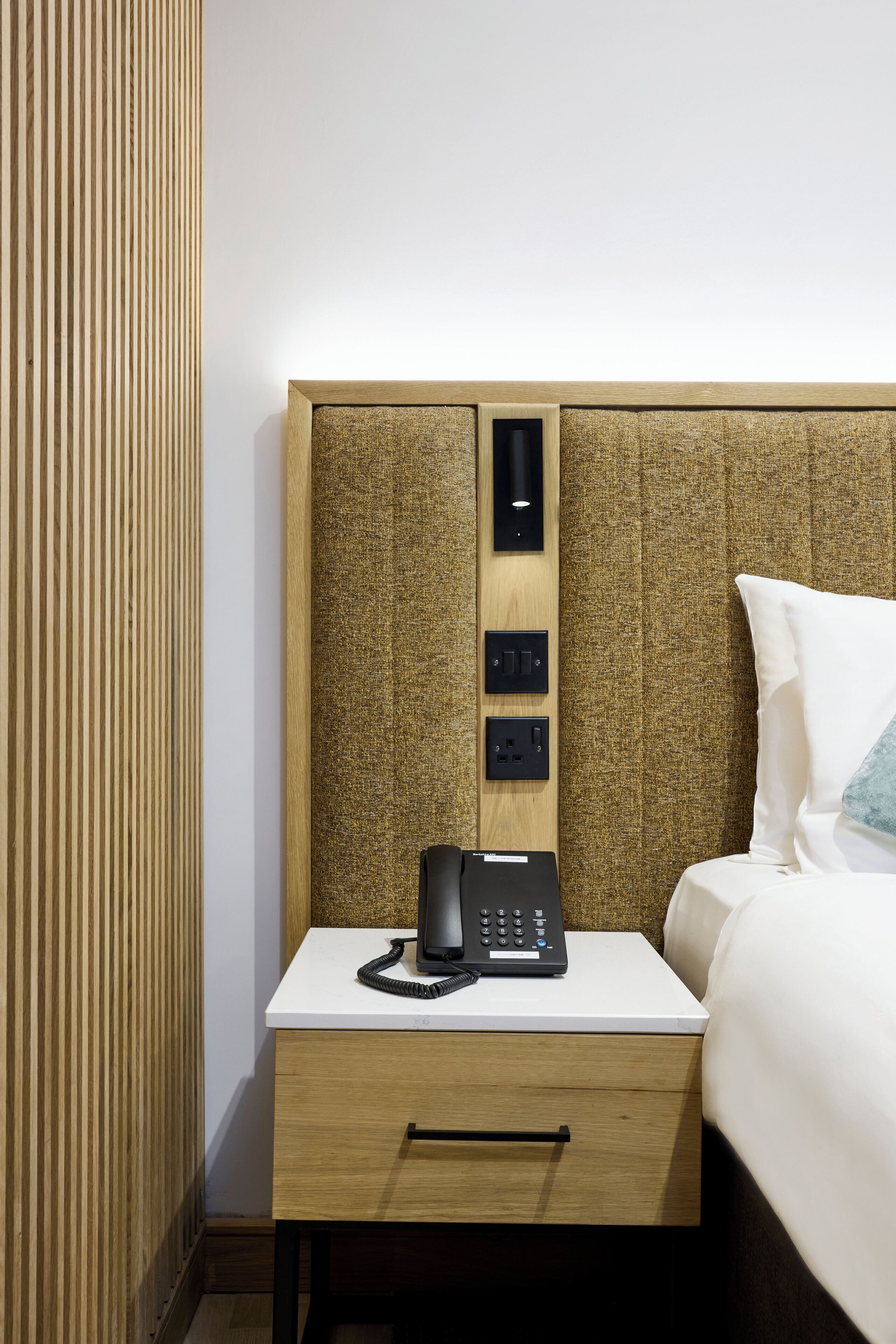 Quarto de hotel com cabeceira de madeira, mesa de cabeceira com telefone e cama branca.