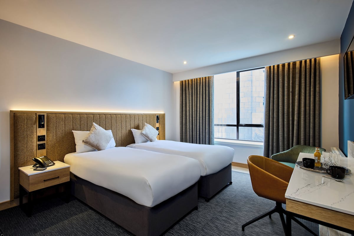Habitación de hotel con dos camas, un escritorio, una silla, un teléfono en la mesita de noche y una ventana con cortinas.