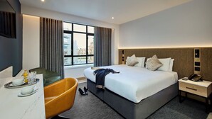 A modern hotel room with a large bed, a desk with a chair, a sofa, and a view of the city through the window. - Handel's Hotel (Dublin)