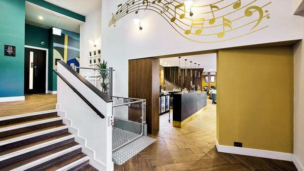 A modern hotel lobby with a staircase, a reception desk, and a wall with musical notes. - Handel's Hotel (Dublin)