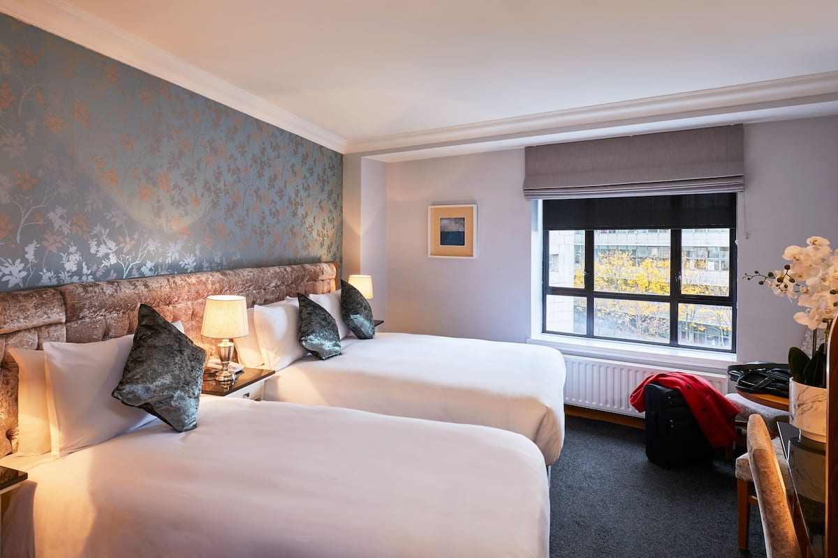 Habitación de hotel con dos camas, ventana con vistas, un radiador y un escritorio con lámpara.