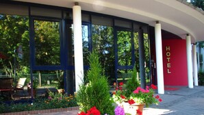 A hotel entrance with large windows, a red wall with the word 'HOTEL' in gold letters, and potted plants with red and purple flowers.