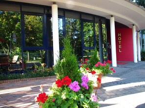 A szálloda bejárata nagy ablakokkal, egy vörös fal arany betűkkel kirajzolt „HOTEL” felirattal, és vörös és lila virágokkal díszített virágtartókban elhelyezett növényekkel.