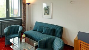 A room with a sofa, armchairs, a glass coffee table, a lamp, a framed picture, and a window with curtains.