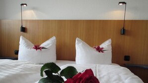 A hotel room with a neatly made bed, two pillows with flowers, a red rose, and two wall-mounted lamps.