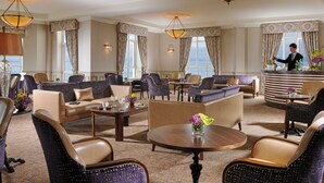 A spacious hotel lobby with multiple seating areas, including sofas and chairs, arranged around a central coffee table. There are large windows providing natural light and a view of the outside.