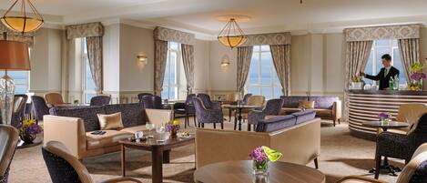 A spacious hotel lobby with multiple seating areas, including sofas and chairs, arranged around a central coffee table. There are large windows providing natural light and a view of the outside.