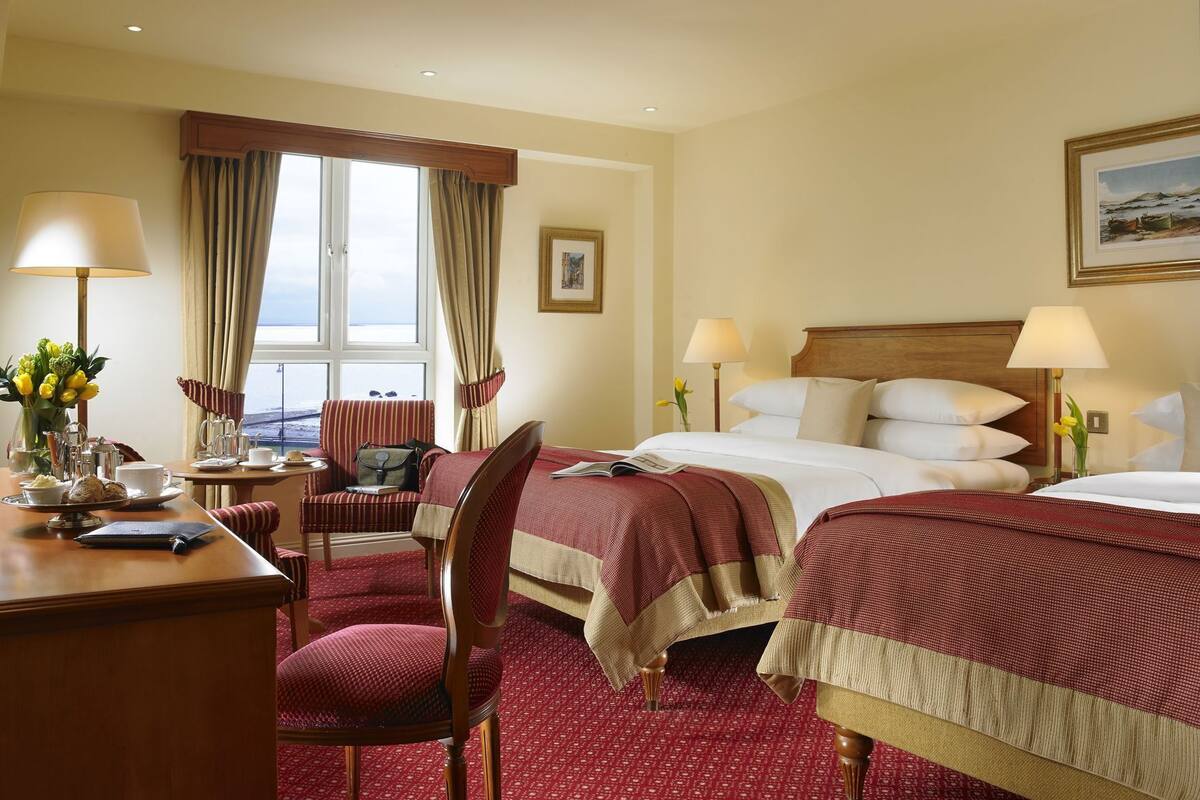 A hotel room with two beds, a desk, a chair, a window with curtains, and a painting on the wall.