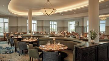 Un vaste espace de restauration dans l’hôtel, offrant différents types d’aménagements, notamment des tables rondes et des banquettes, ainsi qu’un grand lustre.