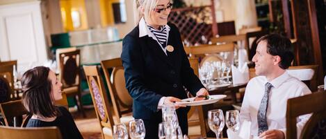 Un'ambientazione di ristorante con un cameriere che serve un piatto a un cliente. I tavoli sono apparecchiati con calici di vino, piatti e posate.