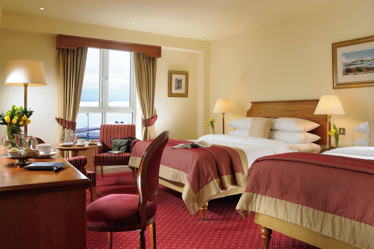 A hotel room with two beds, a desk with a lamp, a chair, and a view of the sea through a window.