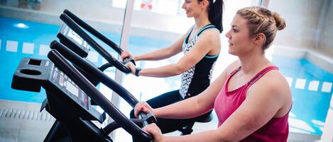 Dos personas usando bicicletas estáticas en un gimnasio con una piscina al fondo.