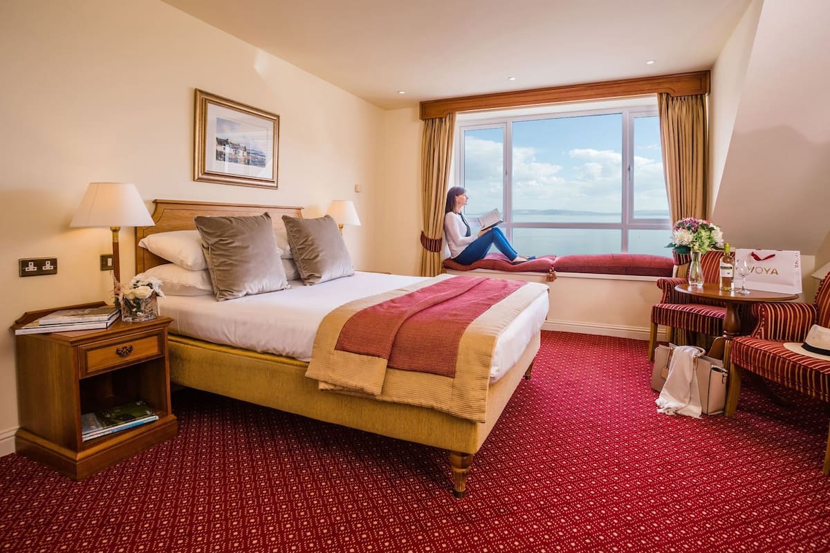 A hotel room with a bed, bedside tables, a window with a view, a seating area with chairs, and a table with a vase and a bag.