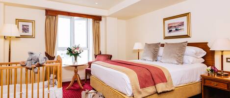 Habitación de hotel con una cama grande, una cuna de madera, un escritorio con lámpara y una ventana con cortinas.