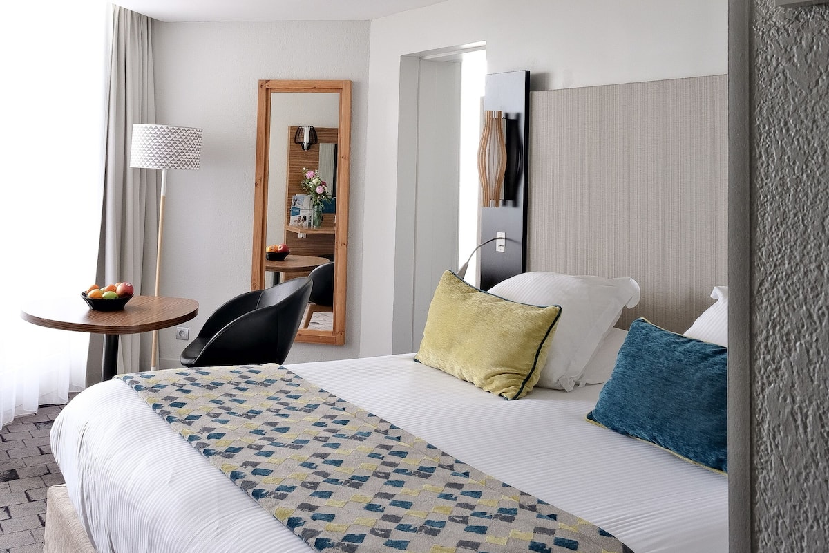 A hotel room with a bed, a desk, a chair, a lamp, and a mirror.