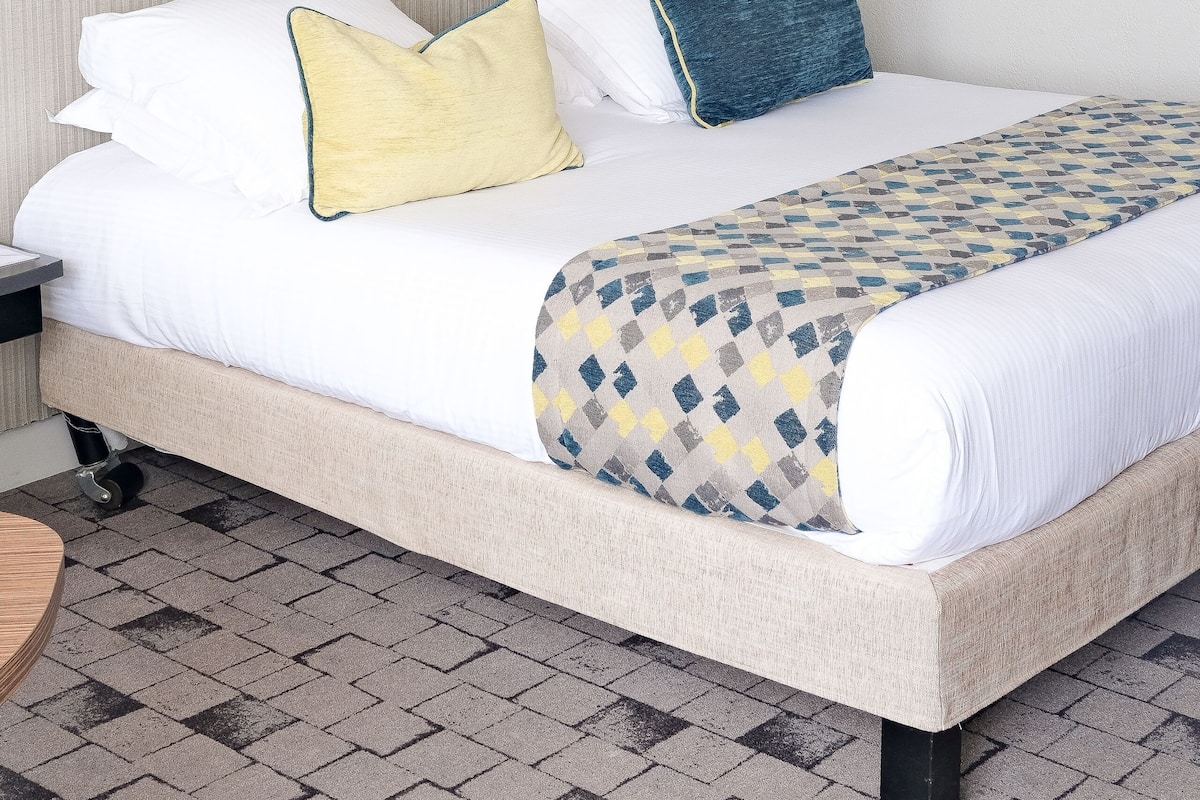 A neatly made bed with white and patterned bedding, a bedside table, and a wall-mounted shelf with decorative items.