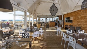 An outdoor dining area with wooden tables and chairs, a bar, and a view of the beach. - Hôtel & Spa - Thalazur Port Camargue (Le Grau-Du-Roi)