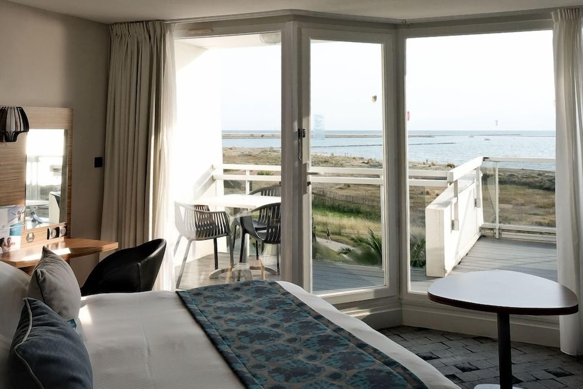Quarto de hotel com cama, escrivaninha, cadeira, mesa e varanda com vista para a praia e oceano.