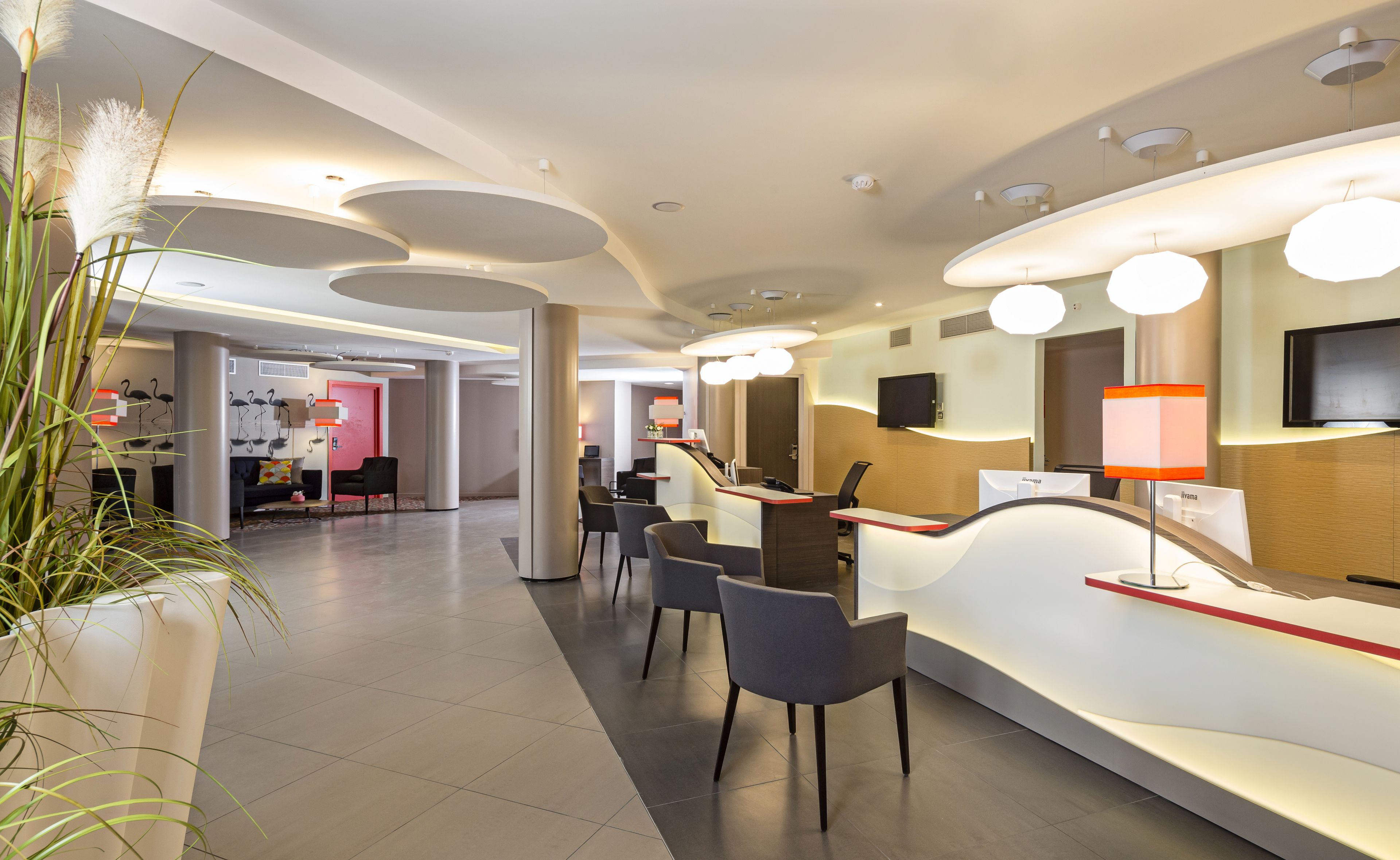 A modern hotel reception area with a curved reception desk, several chairs, and a large potted plant.