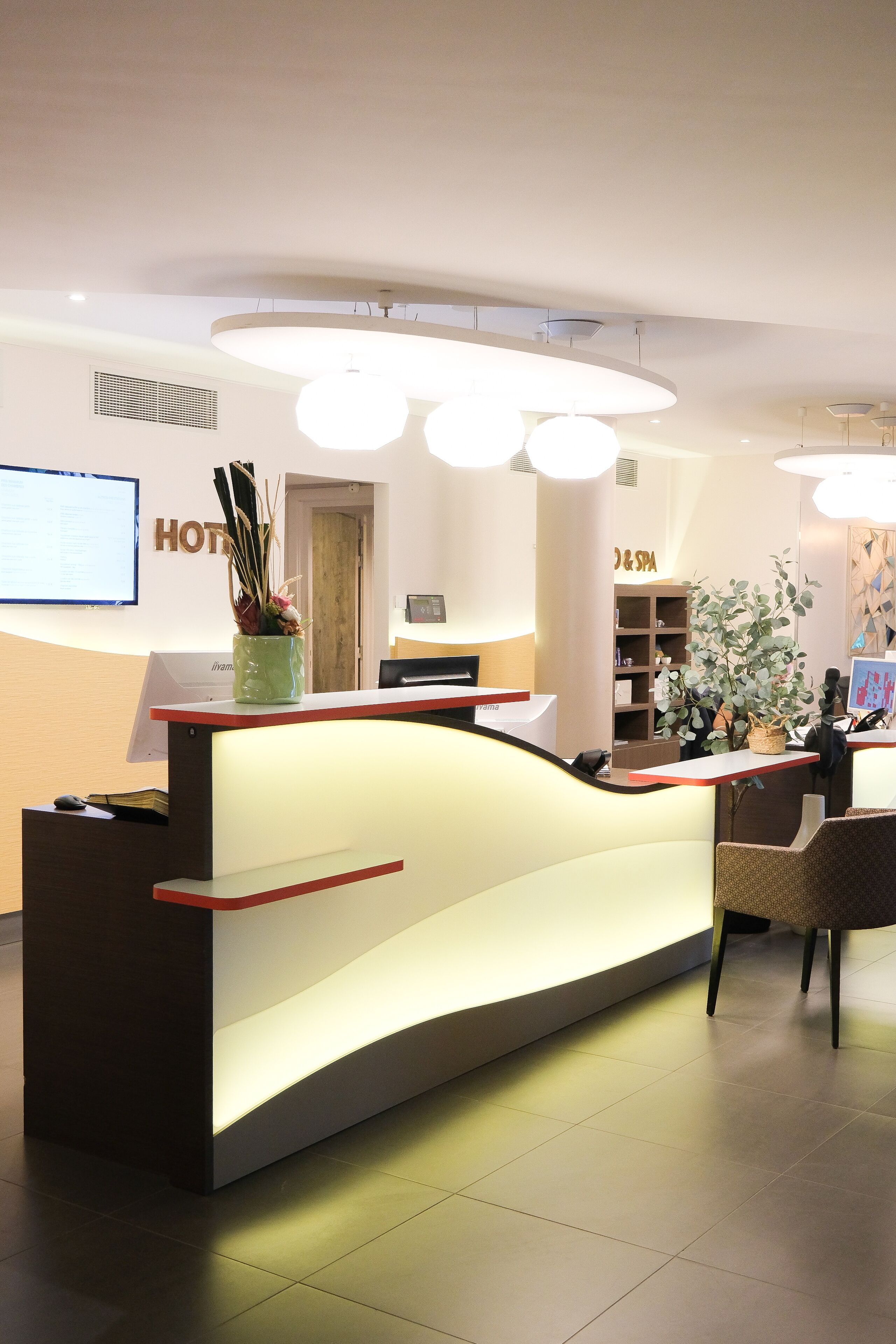 Un mostrador de recepción de hotel con diseño moderno, una pantalla digital y un área de recepción con una planta y un cartel que dice 'HOTEL & SPA'.