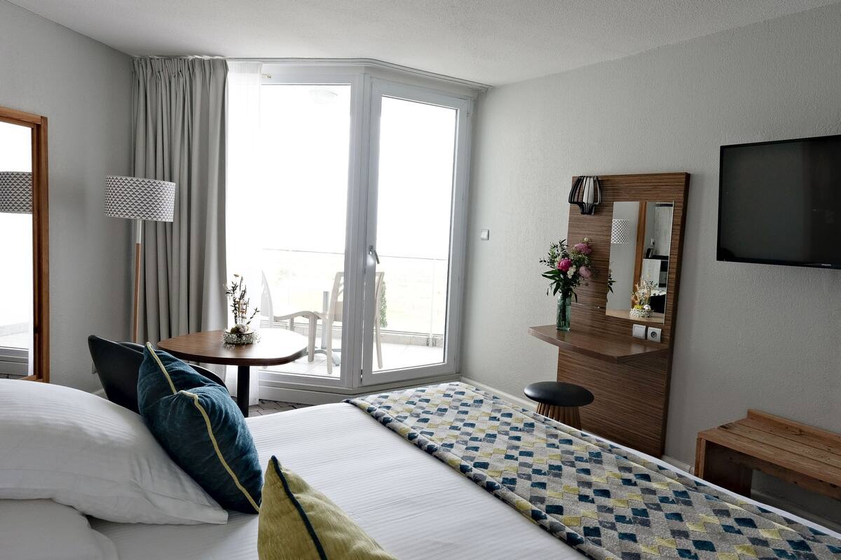 A hotel room with a bed, a desk, a chair, a TV, a mirror, and a balcony with curtains.