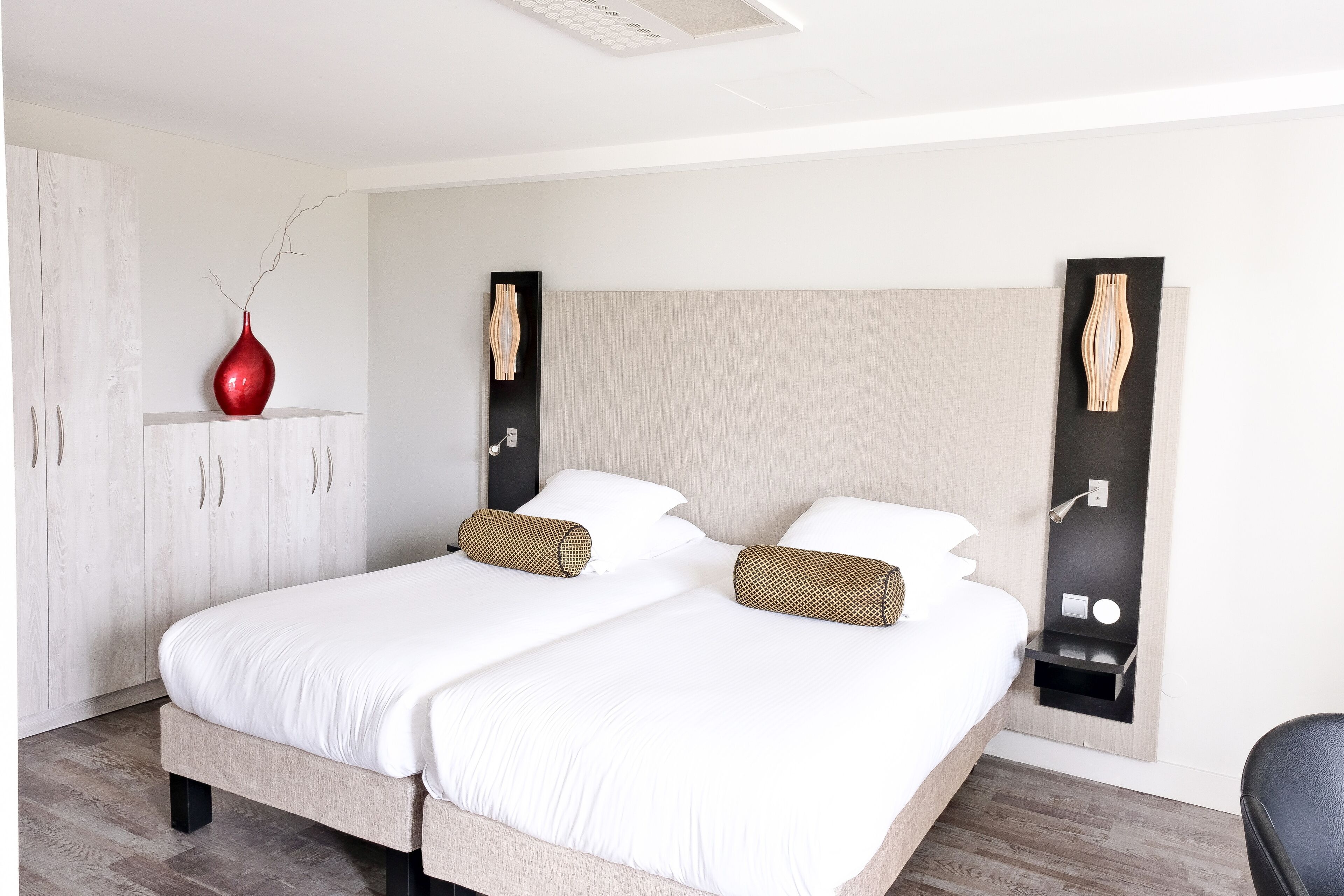 A double bed with white linens, a red vase, and two wall-mounted side tables with wooden sculptures.