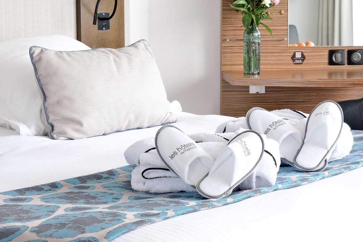 A neatly made bed with a patterned bedspread, a vase with flowers, and slippers on the bed.