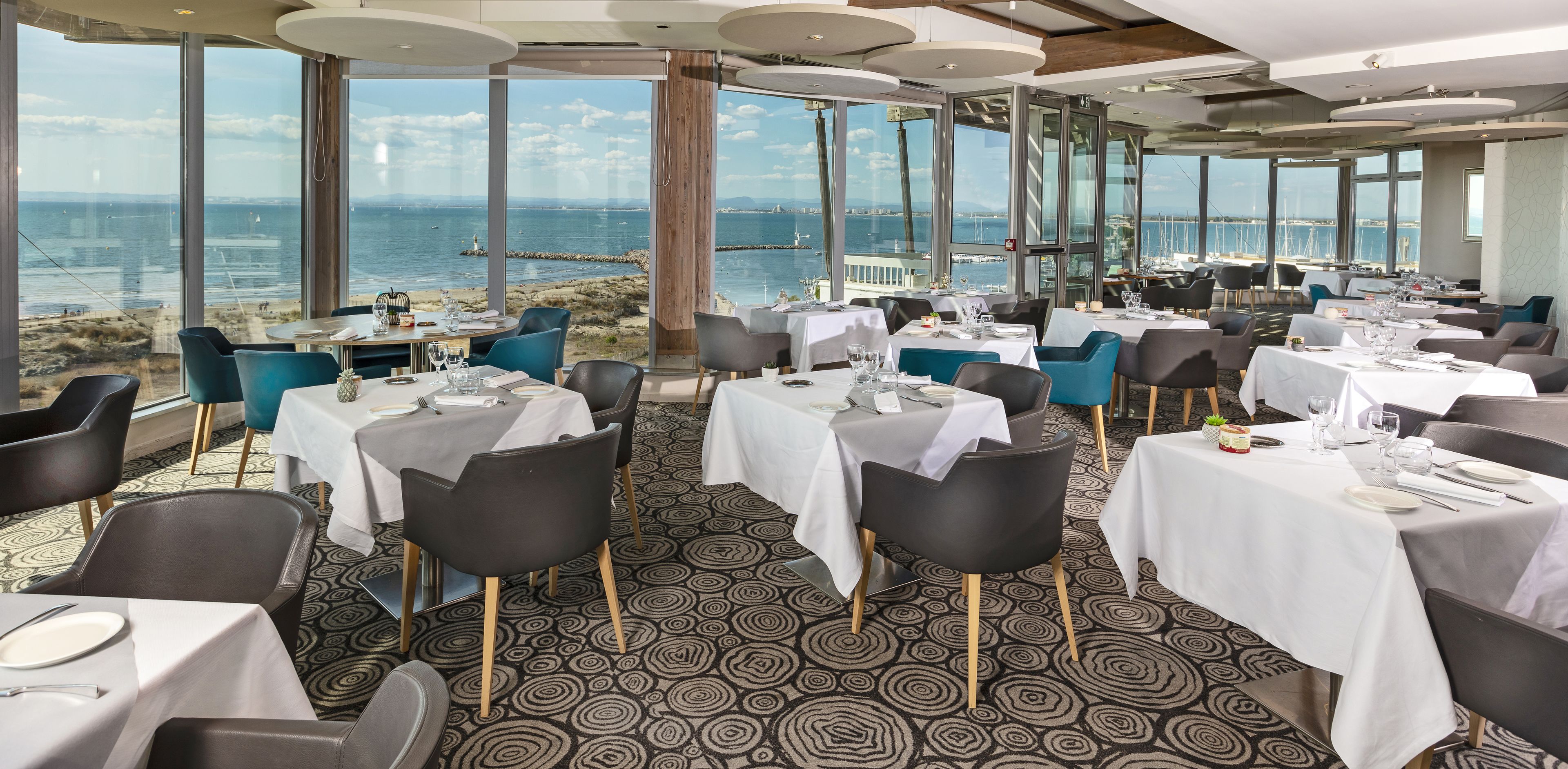 A restaurant with tables set for dining, large windows offering a view of the sea, and a patterned carpet.