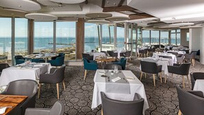 A restaurant with tables set for dining, large windows offering a view of the sea, and a patterned floor. - Hôtel & Spa - Thalazur Port Camargue (Le Grau-Du-Roi)