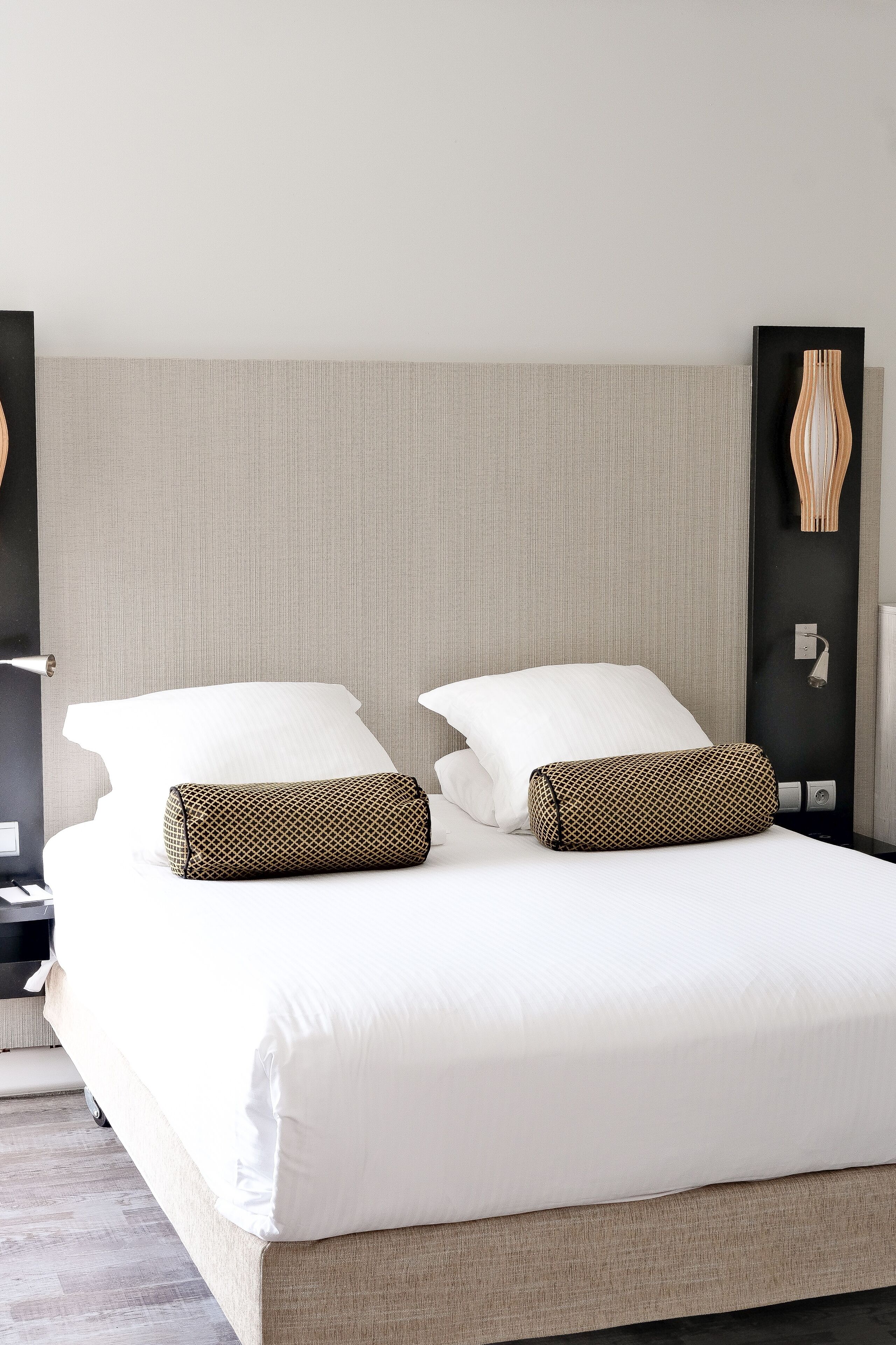 A neatly made double bed with white linens and two patterned pillows, a textured headboard, and a bedside table with a lamp.