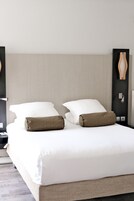 Una cama doble bien hecha con sábanas blancas y dos almohadas estampadas, un cabecero texturizado y una mesita de noche con lámpara.