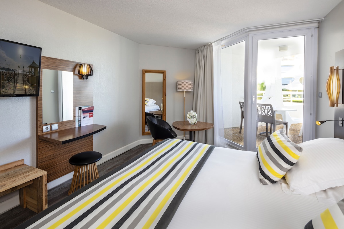 Quarto de hotel com cama, escrivaninha, cadeira, mesinha e vista para o exterior.