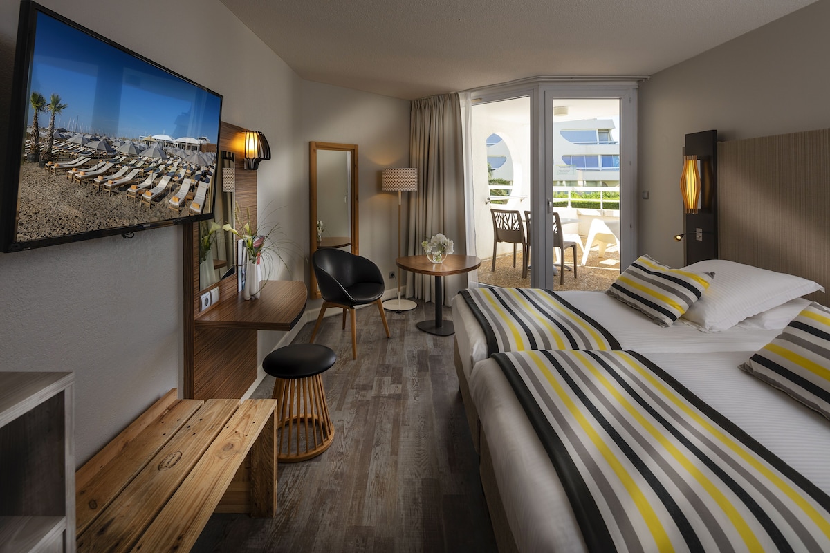 Quarto de hotel com cama, escrivaninha, cadeira, mesa pequena, televisão e vista para a praia.