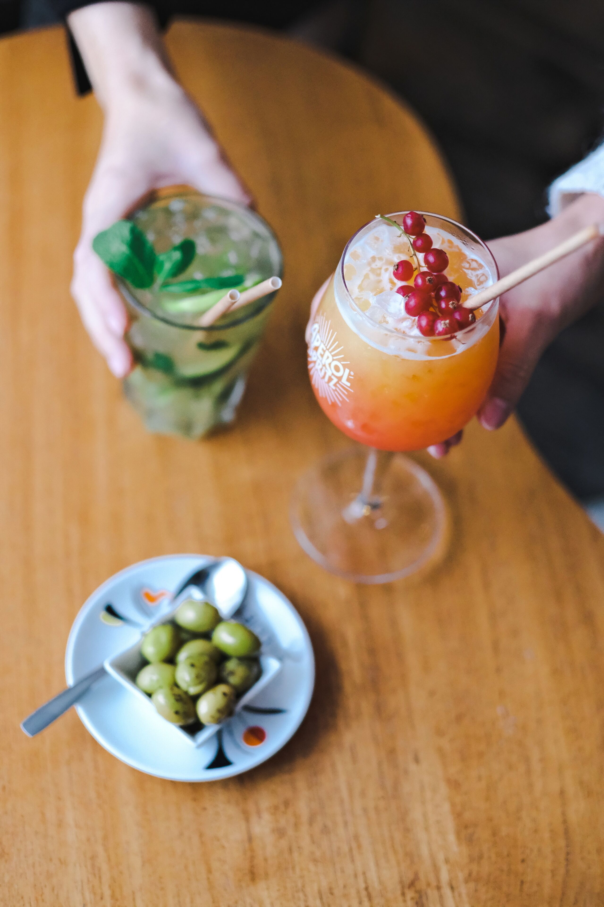 Una mesa de madera con dos bebidas, una en un vaso y otra en un frasco, y un plato pequeño con aceitunas.