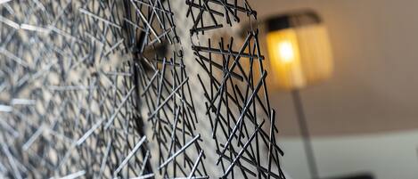 A close-up of a metal mesh structure, possibly part of a wall or partition, with a blurred background featuring a lamp and a chair.