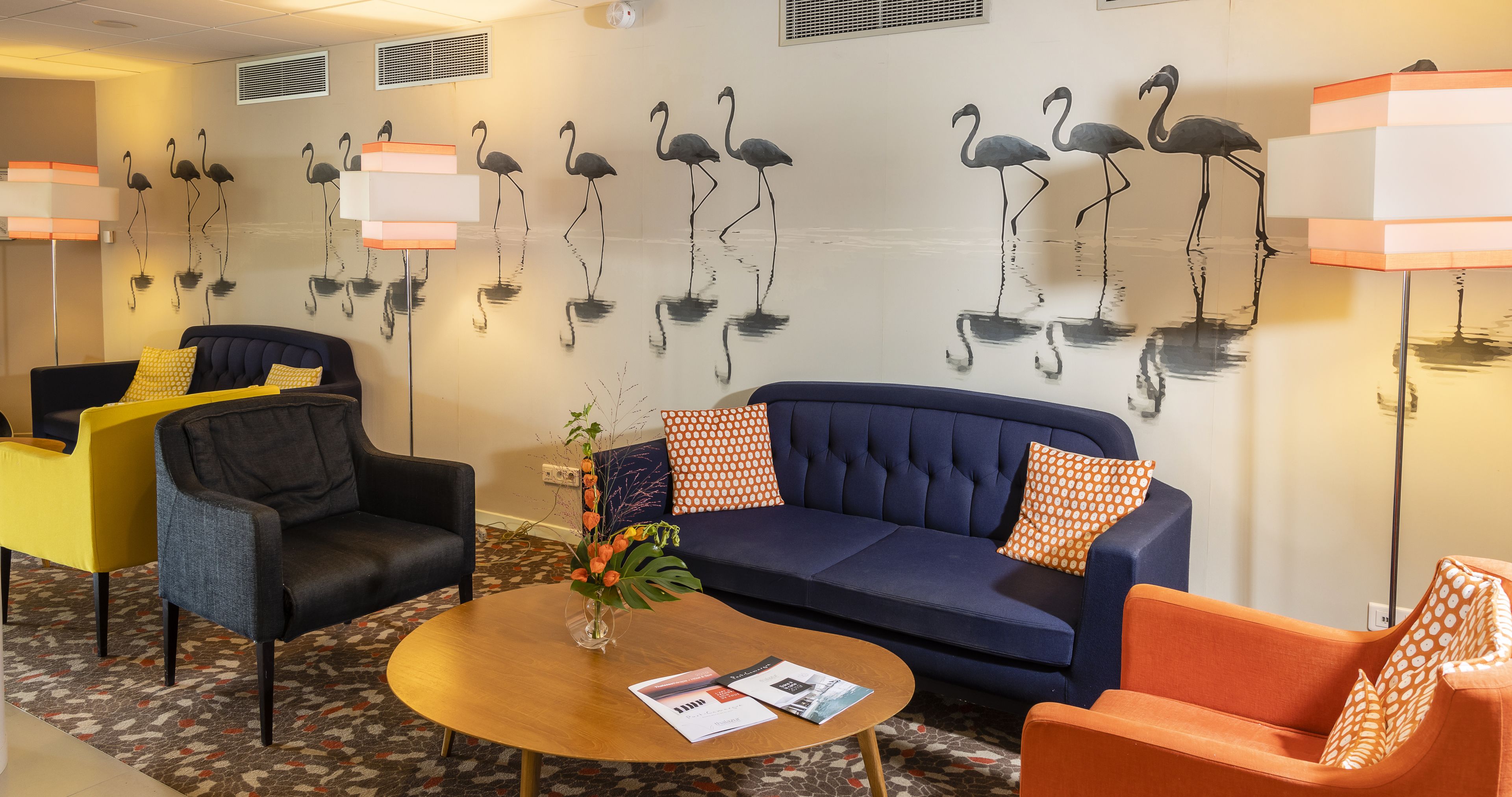 A hotel lobby with a blue sofa, orange and yellow chairs, a round wooden table with a vase and magazines, and a wall mural of flamingos.