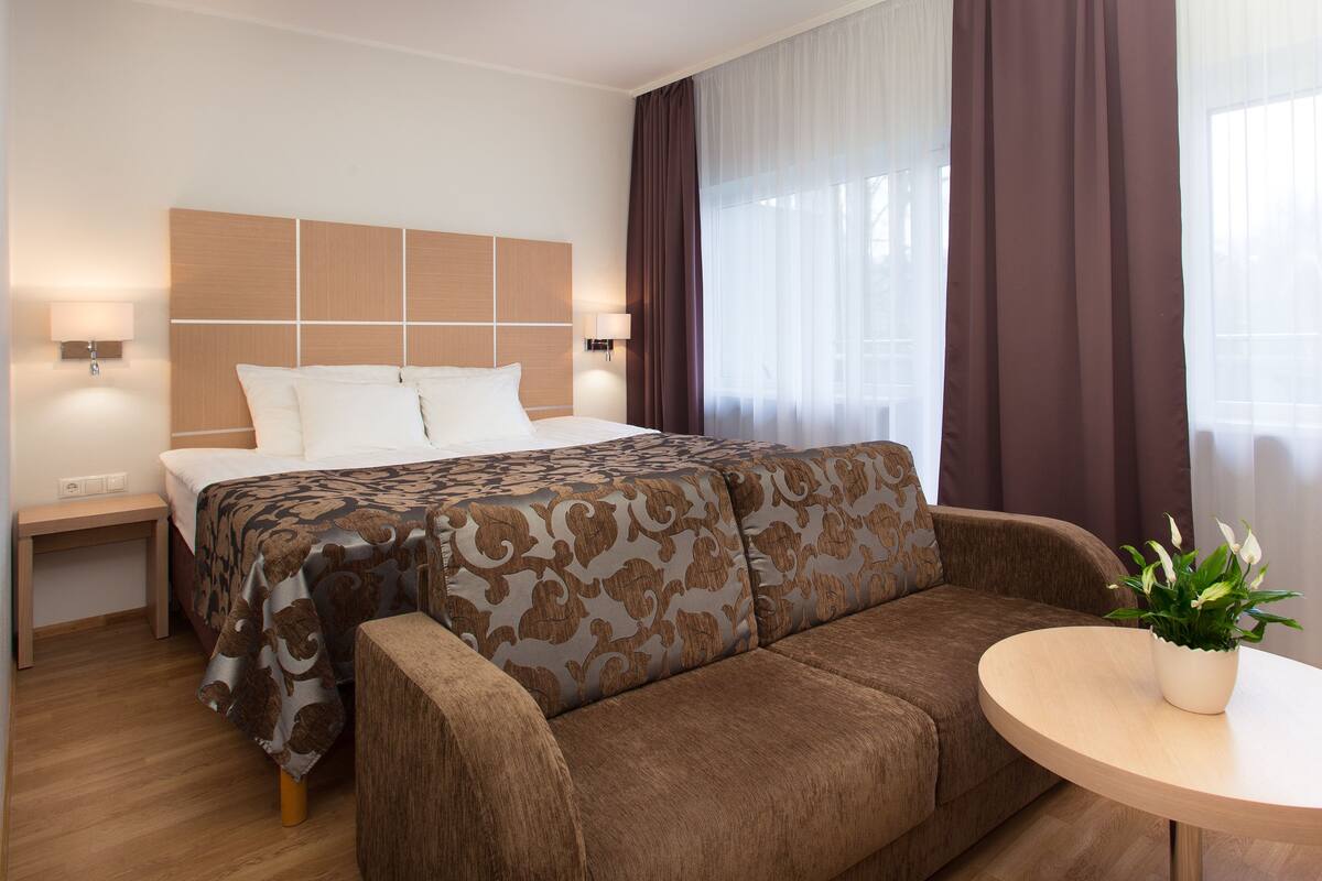Une chambre d’hôtel avec un lit, un canapé, une petite table avec une plante et une fenêtre avec des rideaux.