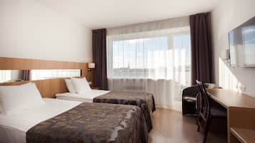 A hotel room with two beds, a desk, a chair, a TV, and a large window with curtains.