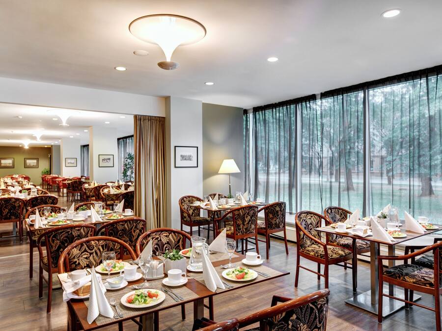 A hotel dining area with multiple tables set for dining, chairs, and a large window with a view of trees.