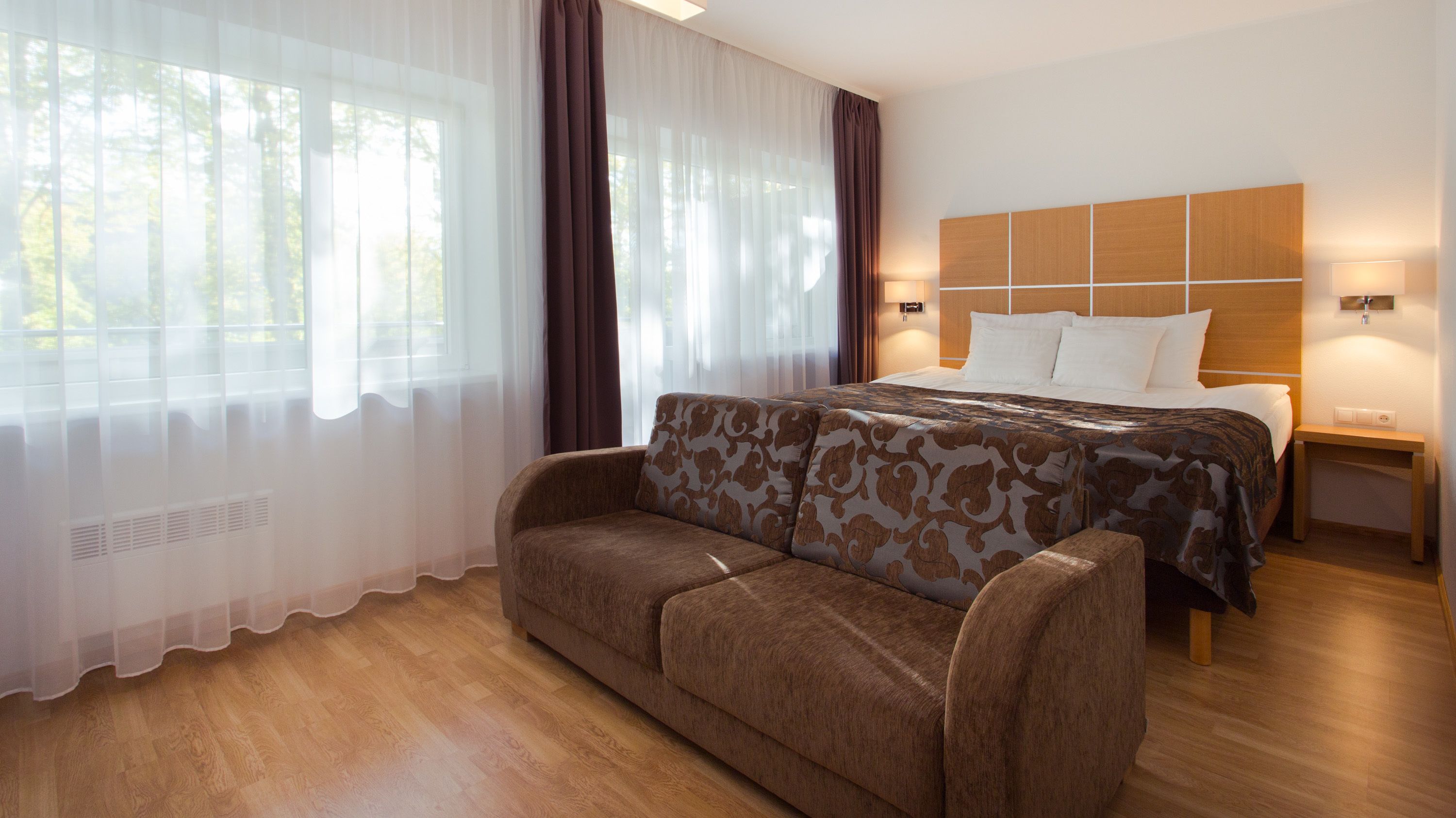 A hotel room with a bed, a sofa, a nightstand, and a window with sheer curtains.