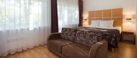 A hotel room with a bed, a sofa, a nightstand, and a window with sheer curtains.