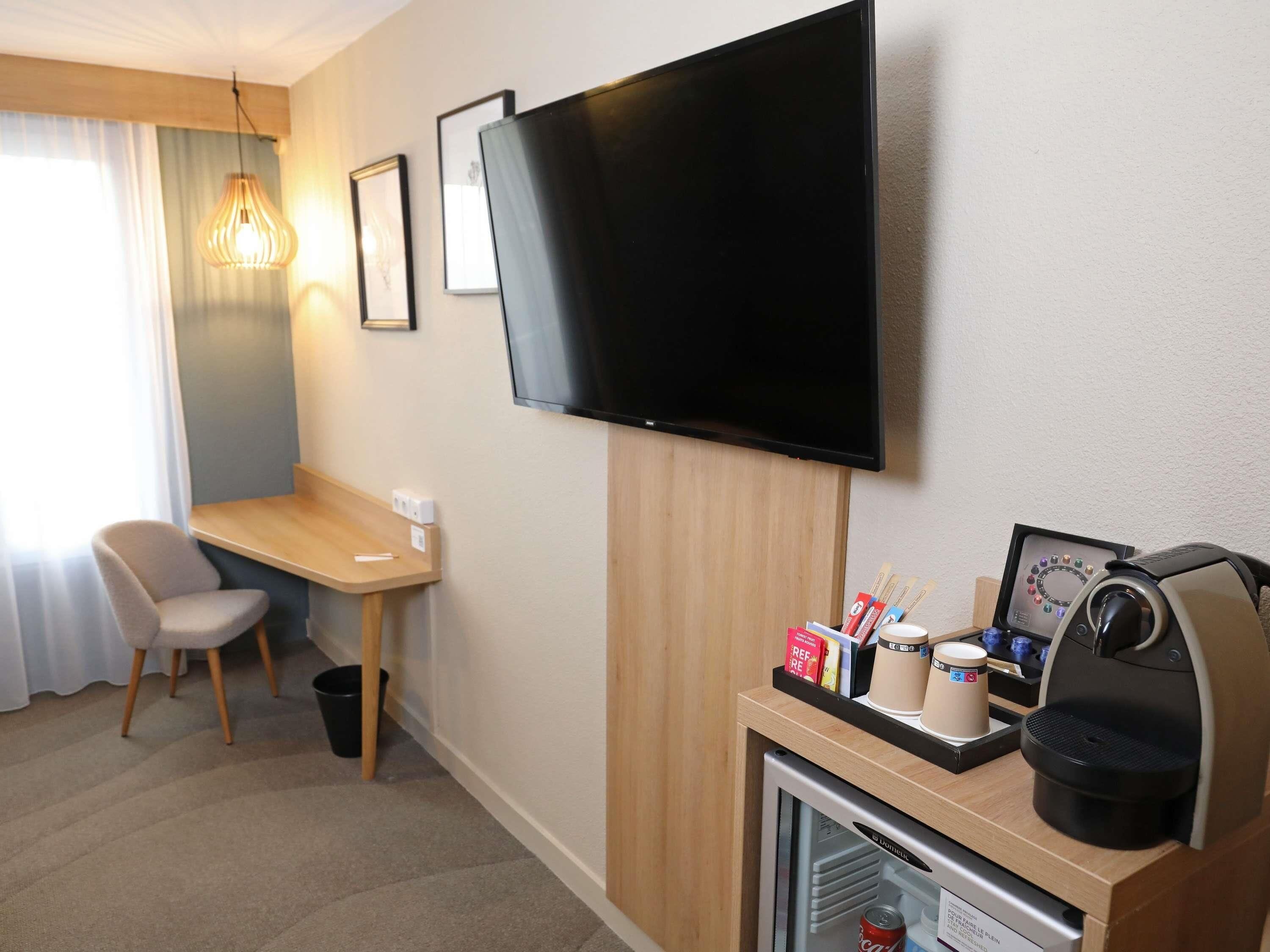 Habitación de hotel con televisor de pantalla plana, un escritorio pequeño, una silla, cafetera y un refrigerador miniatura.