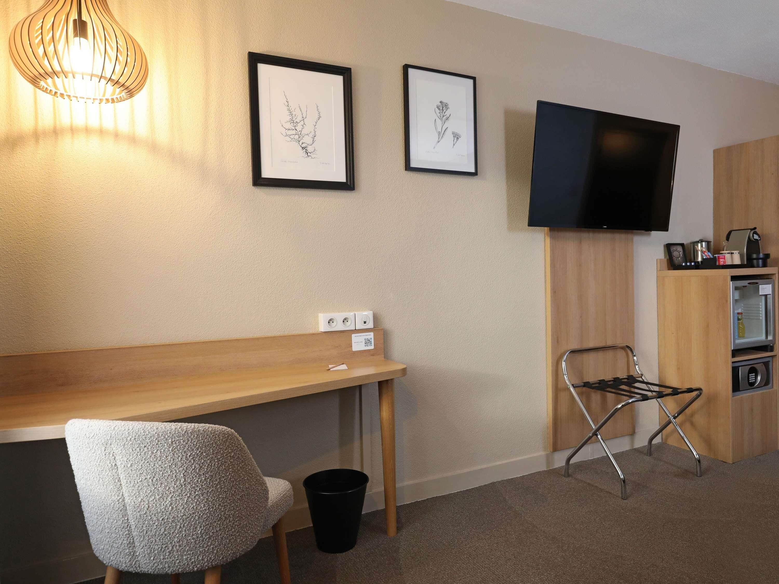 A hotel room with a wooden desk, a chair, a TV mounted on the wall, and a small kitchenette area.