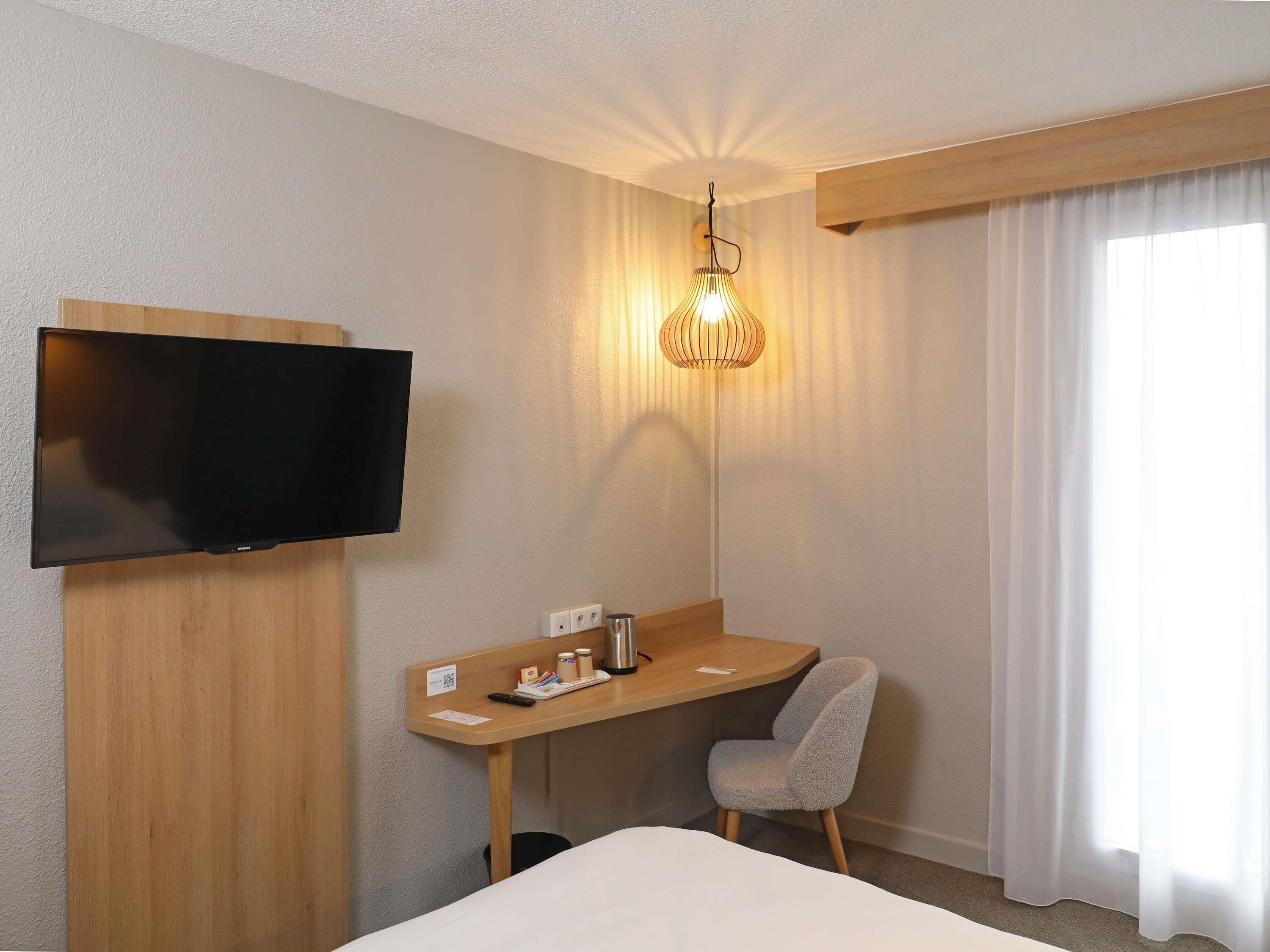 A hotel room with a flat-screen TV mounted on the wall, a wooden desk with a chair, a small table with toiletries, and a large bed.