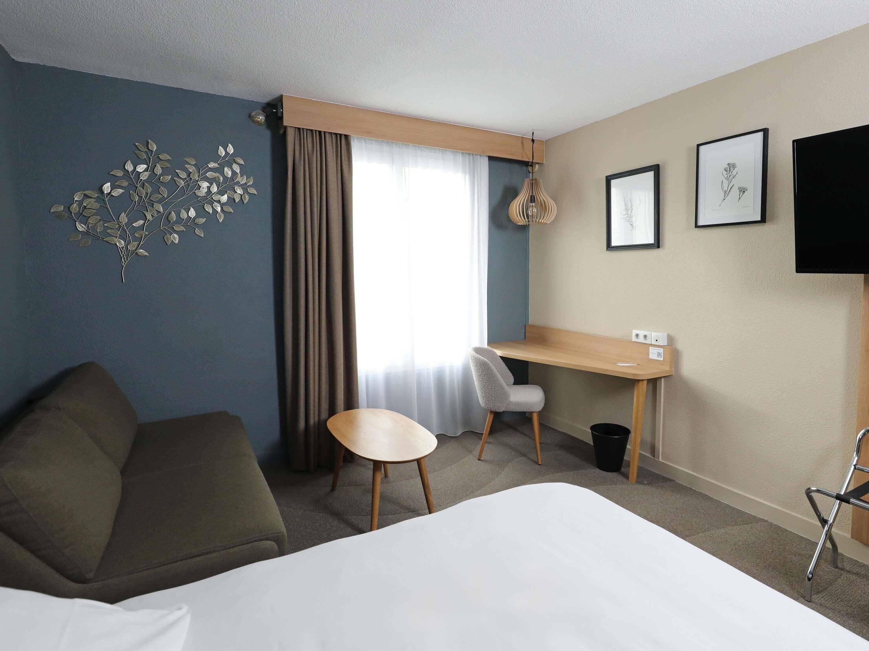 A hotel room with a bed, a desk, a chair, a sofa, a window with curtains, a wall-mounted TV, and a wall art piece.