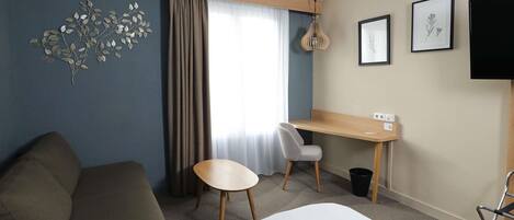 Habitación de hotel con cama, escritorio, silla, sofá, ventana con cortinas, televisor montado en la pared y una obra de arte en la pared.